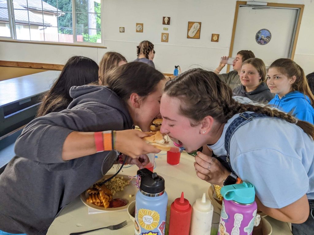 two girls leaning over a cafeteria table, feeding each other hamburgers