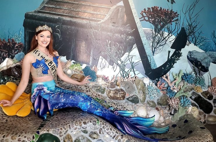 a woman dressed as a mermaid sitting in front of a sunken ship backdrop
