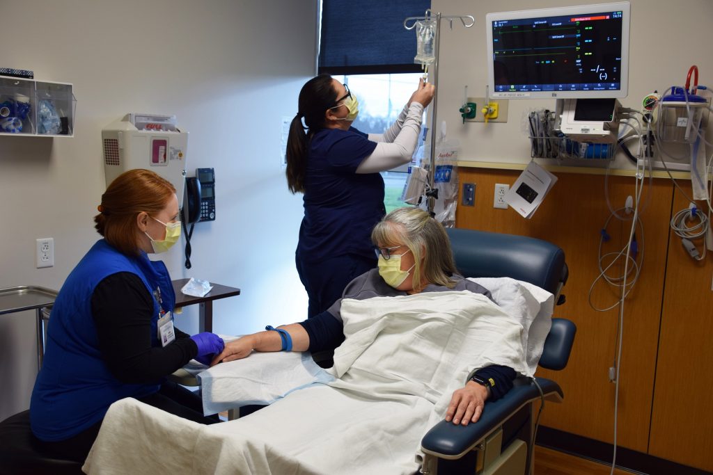 a person in a chair with a white blanket getting an IV infusion with a nurse sitting in front of her and one standing behind her