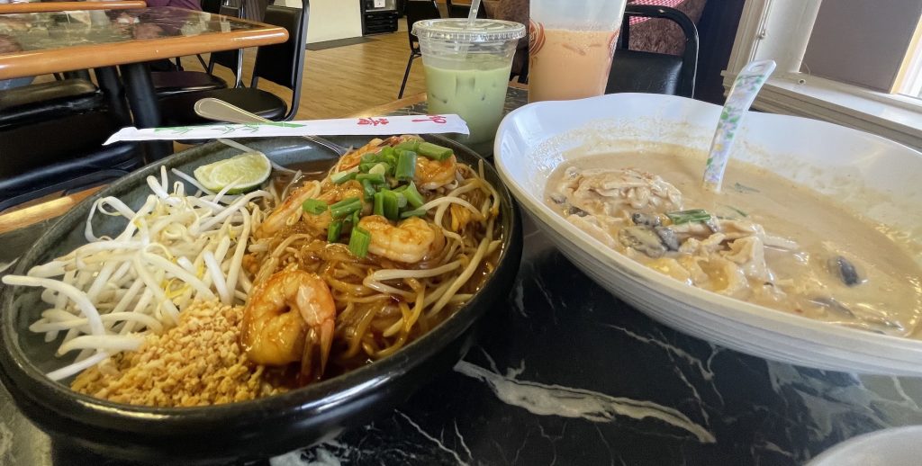 plate of pad thai, bowl of some type of thai soup with two boba tea drinks in the background
