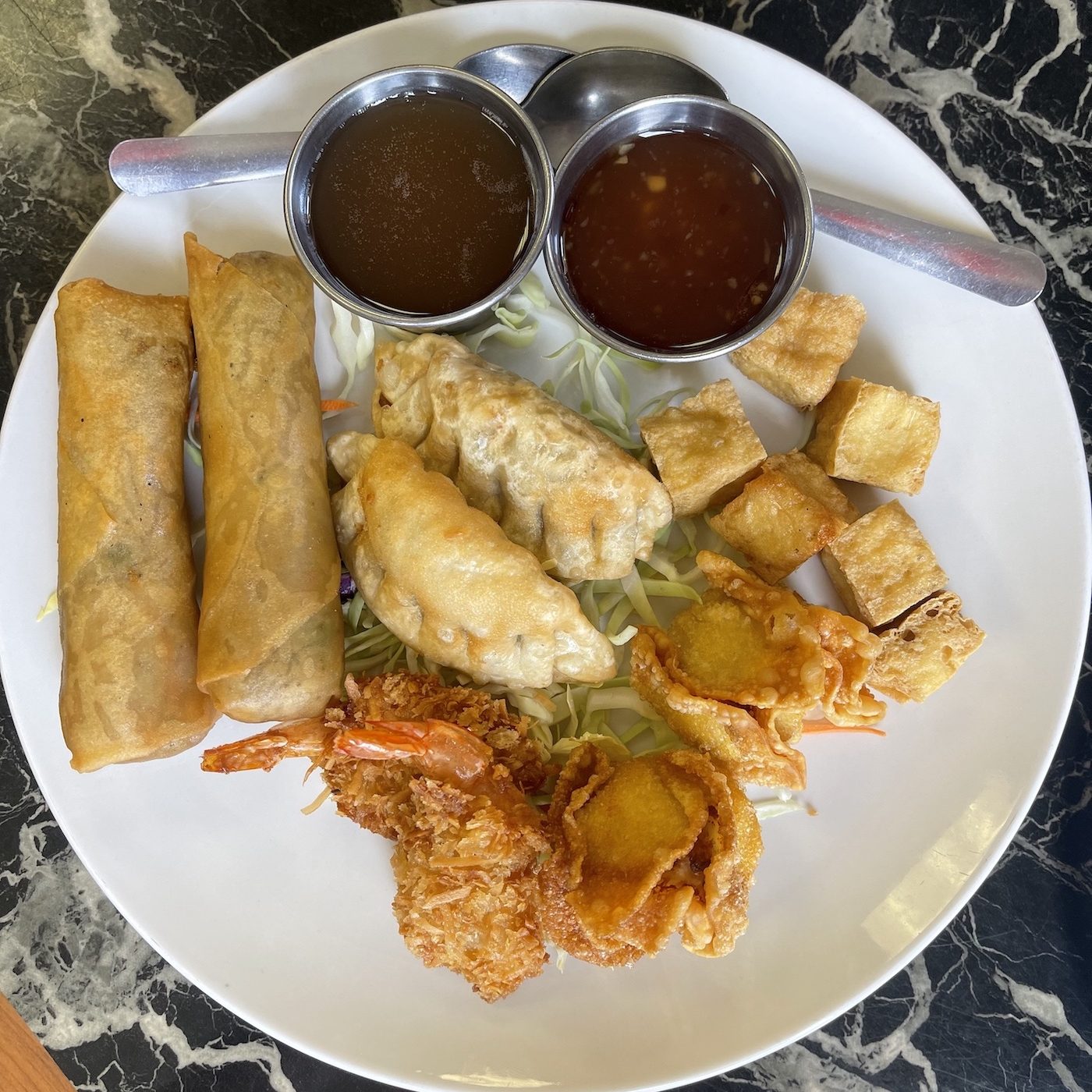 white plate with fried veggie spring roll, chicken cheese rangoon, coconut prawns, pot stickers and four fried tofu. with two small cups of sauce.