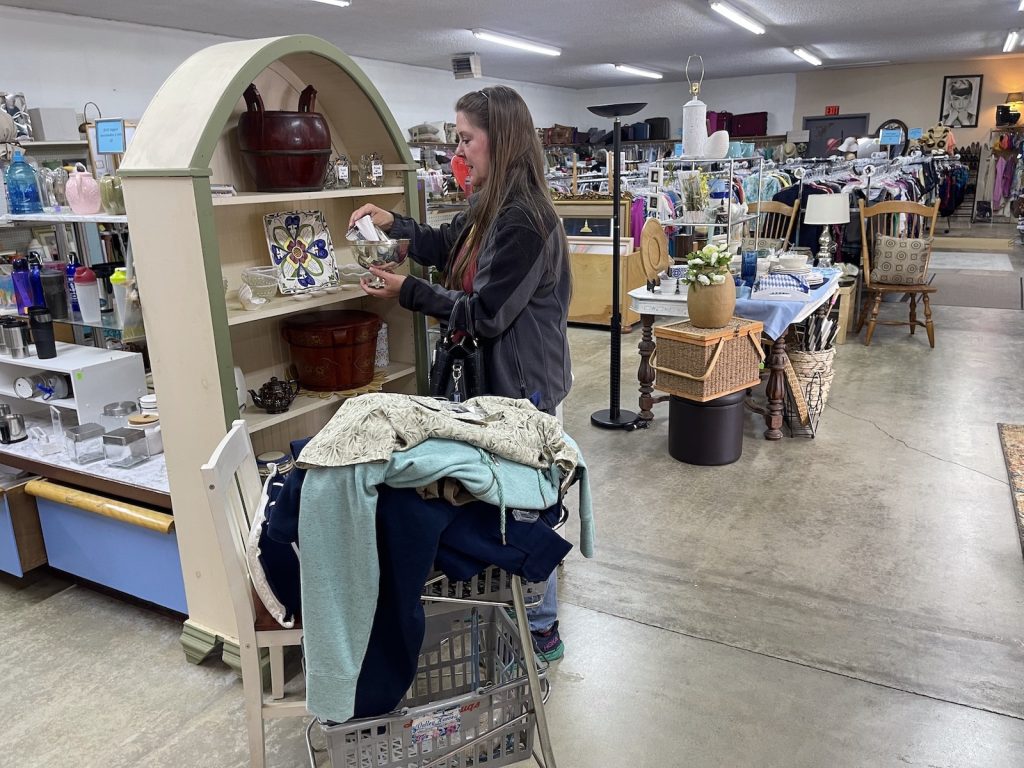 A woman with a full shopping cart of clothes looks at a bowl on a shelf