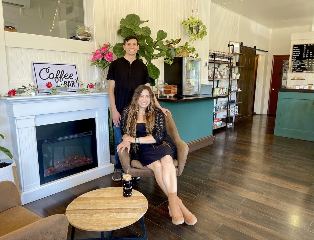 Brass Hub Coffeebar Owners Krissi Brunoe and Mario Barajas pose, Krissi in a chair and Mario behind her, in their coffee shop