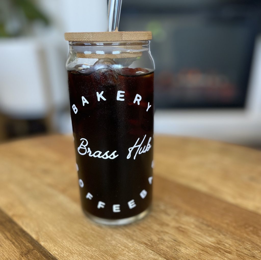 Iced coffee in a glass with a lid and a glass straw. Glass says, "Bakery. Brass Hub Coffeebar'