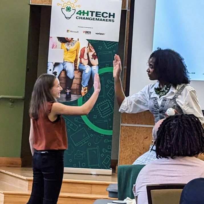 Lucy in a large audtior, giving a woman a high-five, a banner behind them says '4H Tech changemakers'