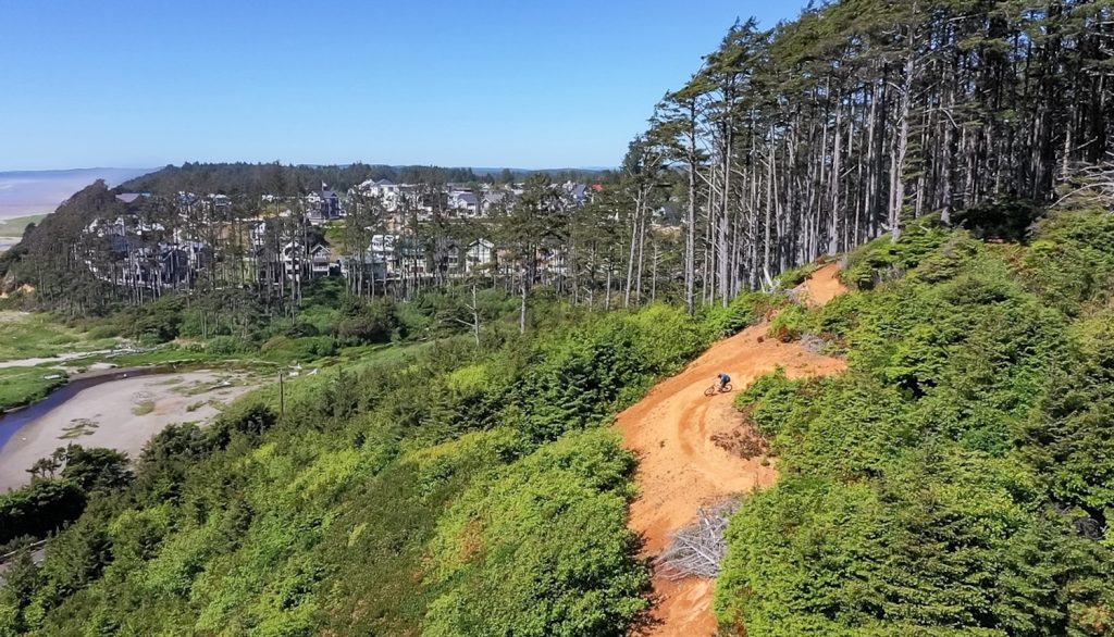 a steep mountain bike trail with a biker on it going down a tree- and bush-lined hill with Seabrook in the distance