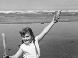 black-and-white photo of a young girl kneeling in the wet sand with a clam shovel in one hand and a clam raised up in the other. A bucket is next to her.