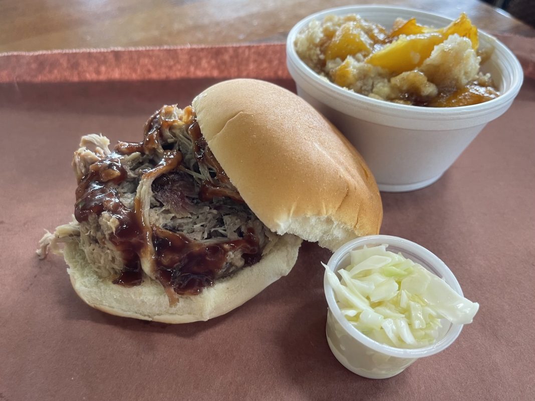 pulled pork sandwich on a tray with a cup of coleslaw and peach cobbler next to it