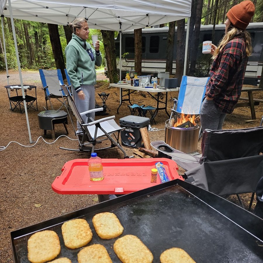 People standing around a campsite with a pop up tent, food and drinks on tables and an RV in the background in the woods in Grays Harbor