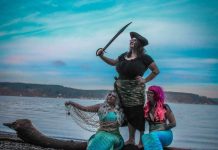 two women dressed as mermaids and one as a pirate pose on a beach for a photo