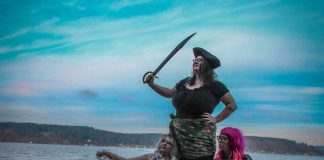 two women dressed as mermaids and one as a pirate pose on a beach for a photo