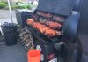 a huge black trailer grill full of meat cooking on it, firewood is stacked beneath it