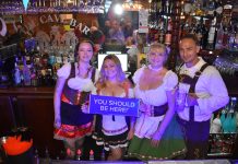 four people dressed in lederhosen for Oktoberfest at The Cave Bar, one woman holds a sign that says 'you should be here'